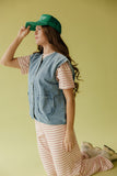 Woman wearing a green cap, denim vest, and striped shirt against a yellow background