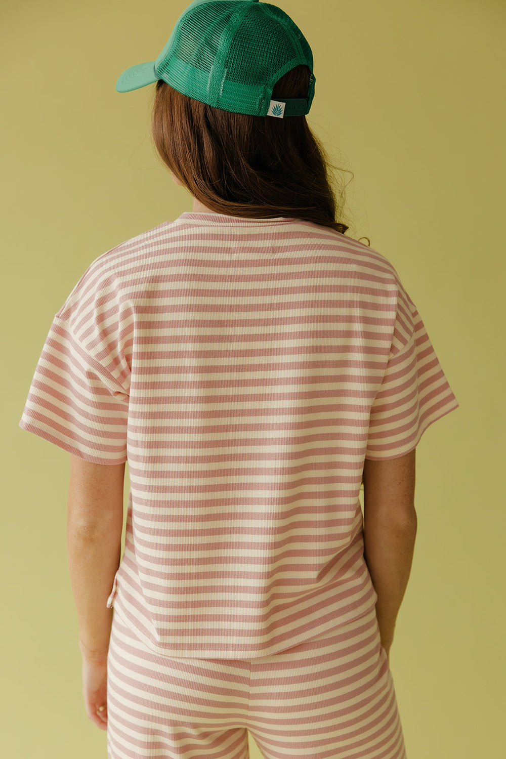 Person wearing a green cap and pink striped shirt and pants on a yellow background