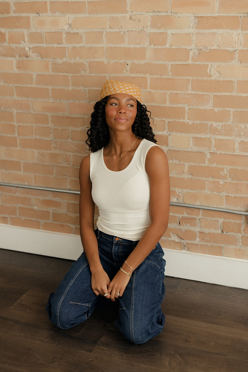 Woman kneeling in front of a brick wall wearing a white tank top and blue jeans.