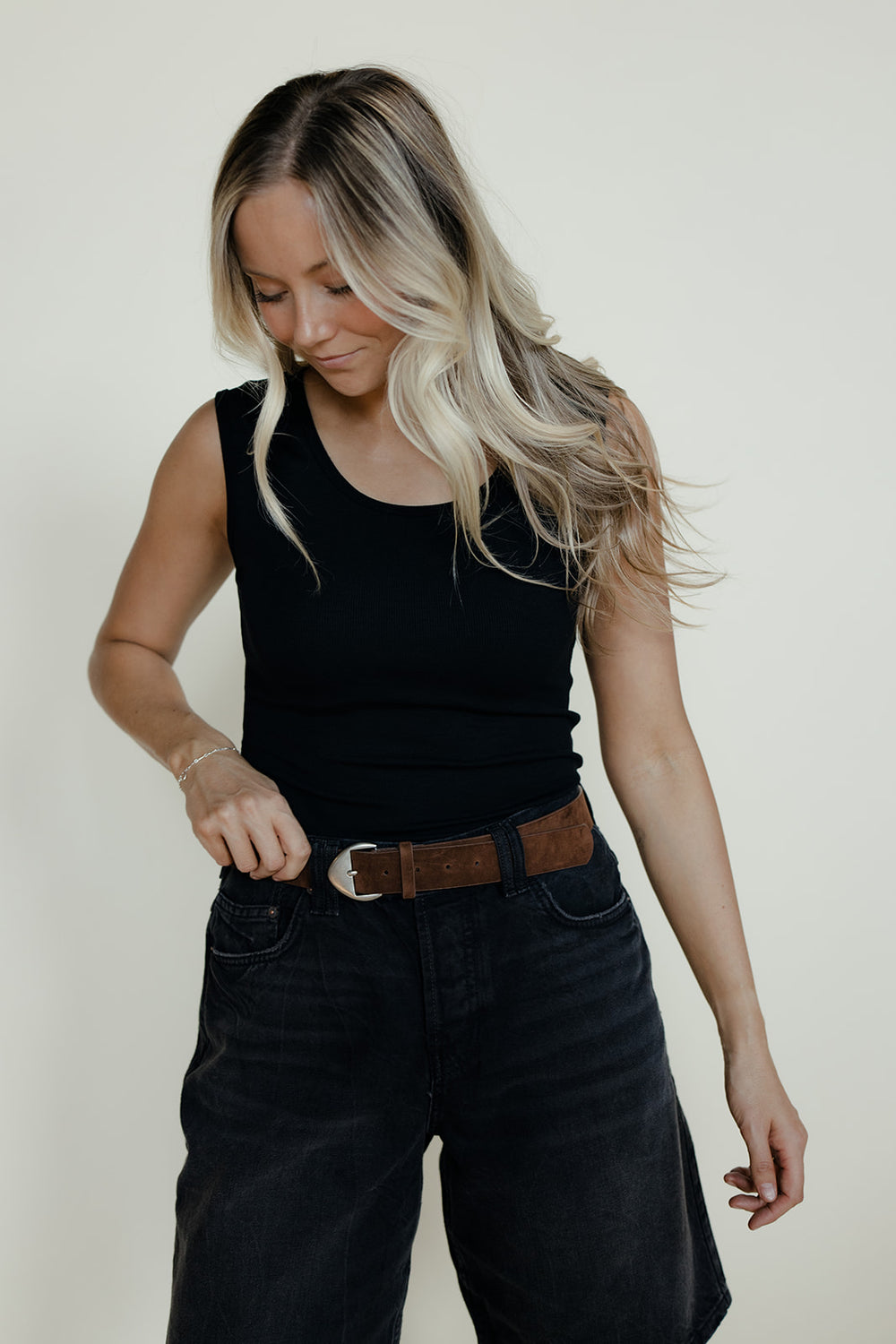 Woman wearing a black sleeveless top and black pants with a brown belt against a plain background