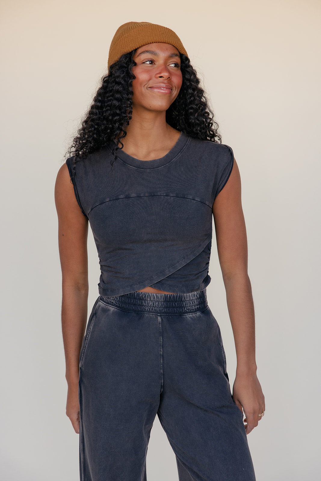 Woman wearing a dark sleeveless top and jeans with a beige cap against a plain background