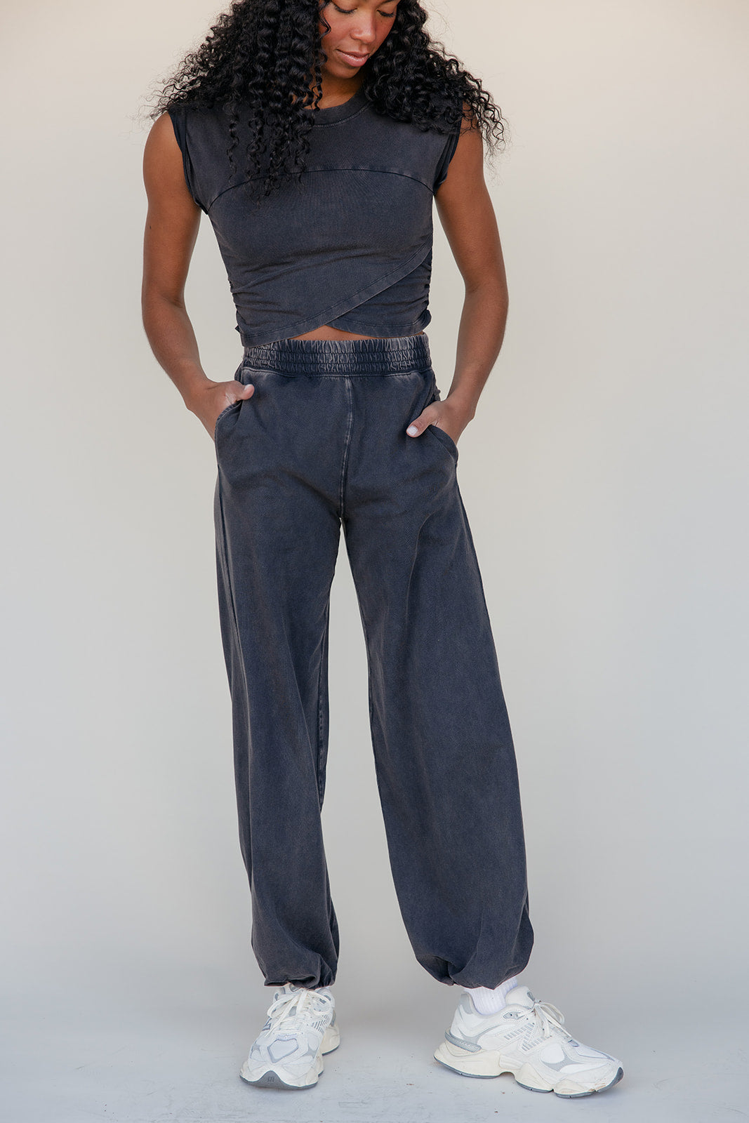 Person wearing a sleeveless dark top and high-waisted dark jeans with white sneakers on a plain background