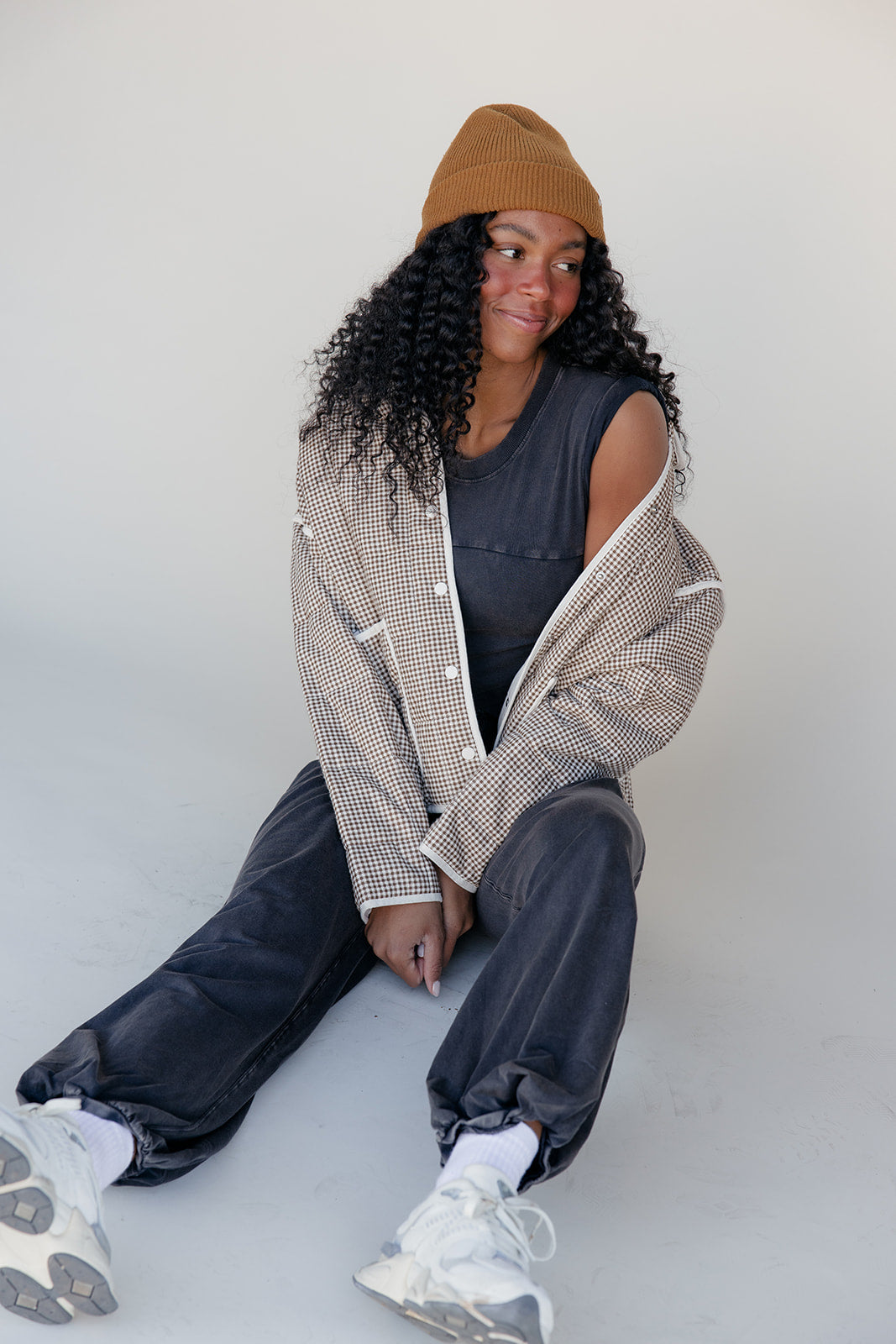 Person wearing a checkered jacket, black sleeveless top, and dark pants sitting against a white background.