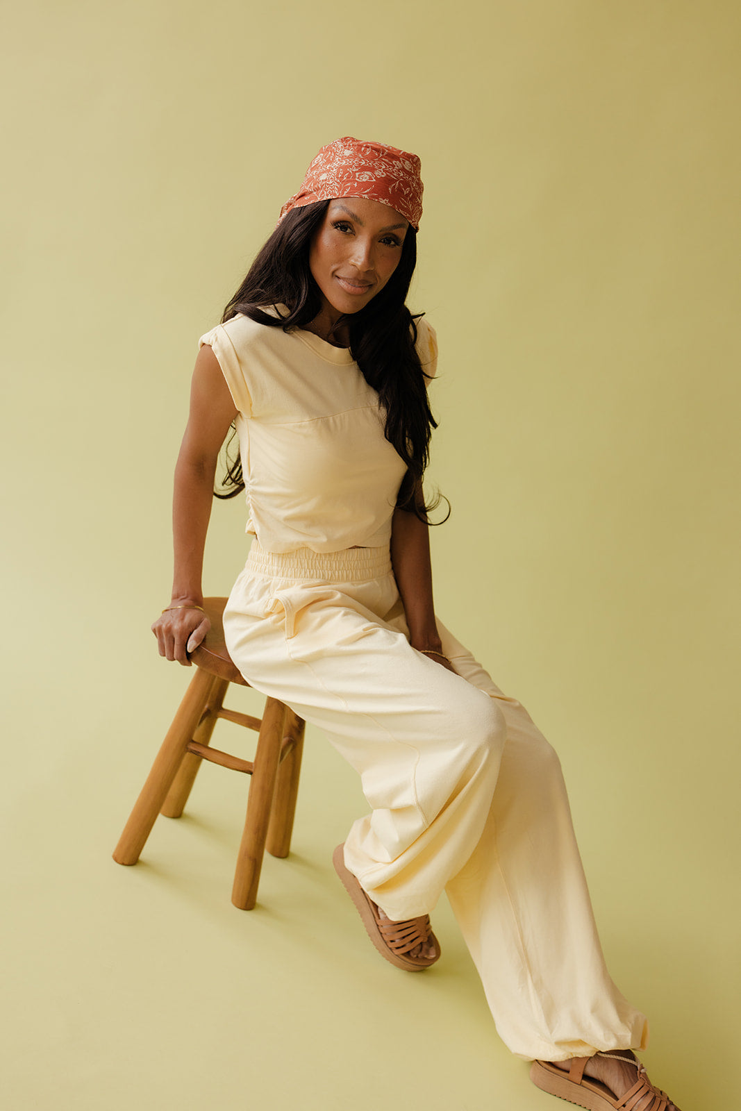 Woman in a beige outfit sitting on a wooden stool against a light green background