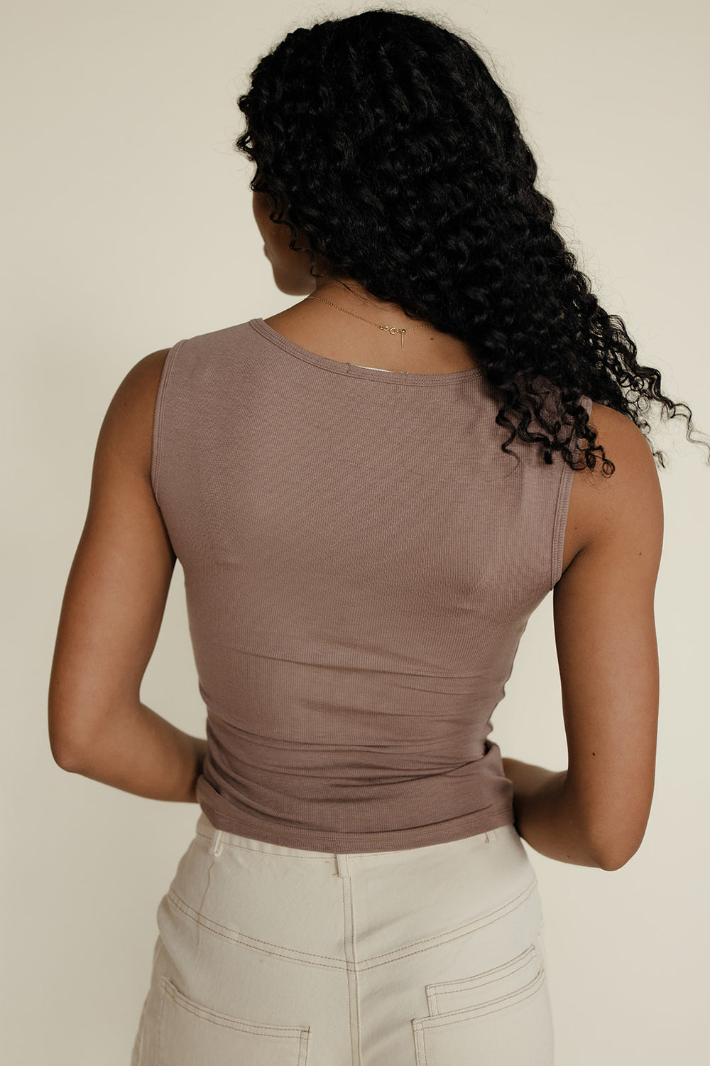 Person wearing a brown sleeveless top and beige pants against a plain background