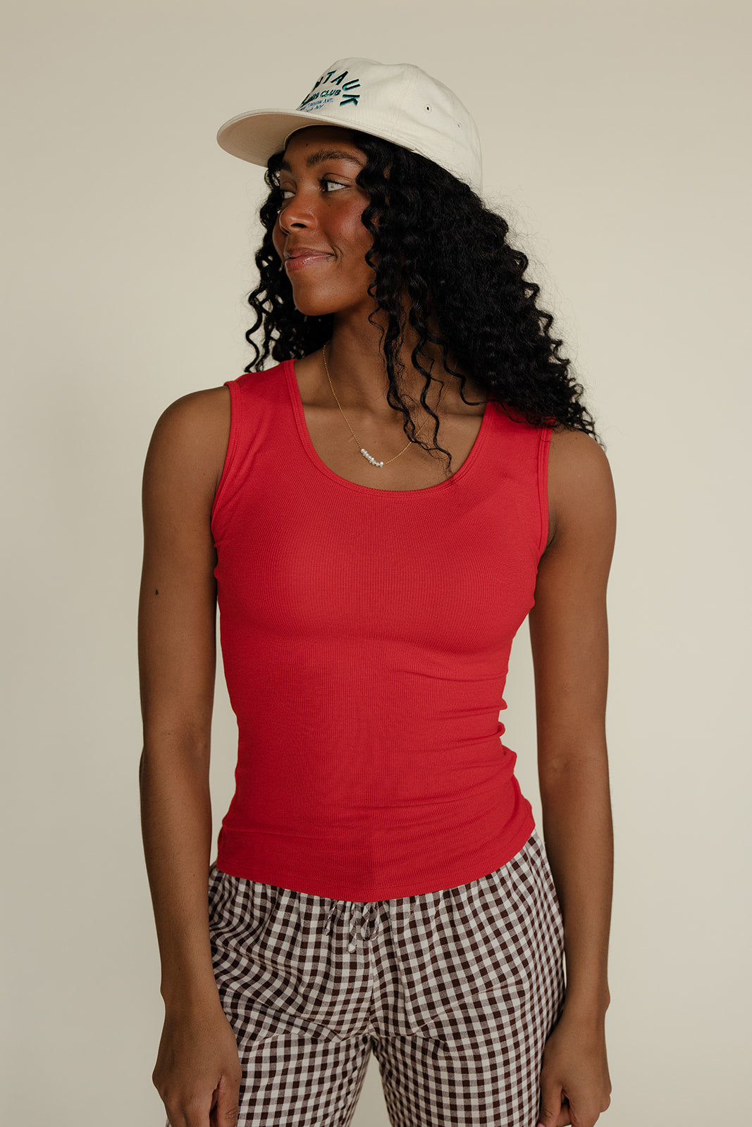 Woman wearing a red tank top and checkered pants with a white cap against a plain background