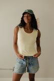 Woman wearing a beige tank top and denim shorts with a green cap indoors.