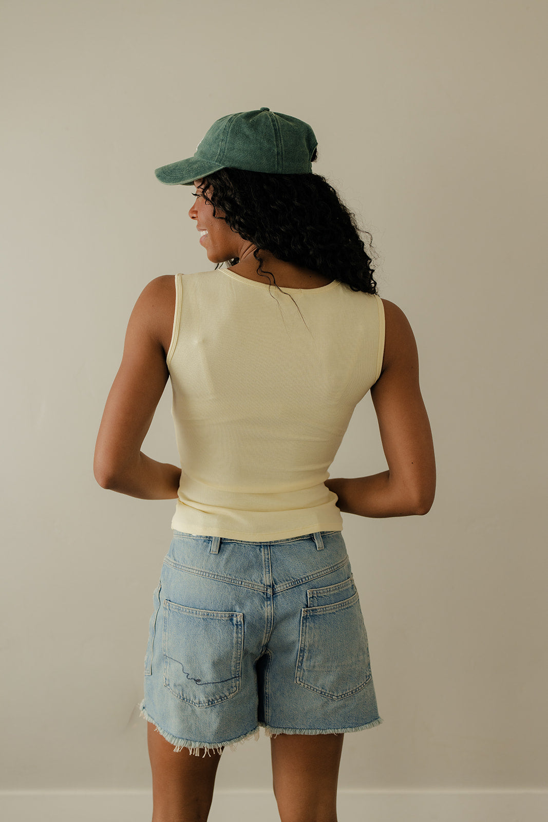 Person wearing a beige sleeveless top, denim shorts, and a green cap against a plain background