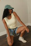 Woman wearing a green cap, white tank top, denim shorts, and white sneakers sitting on the floor.