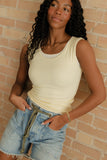 Woman wearing a beige tank top and denim shorts against a brick wall.
