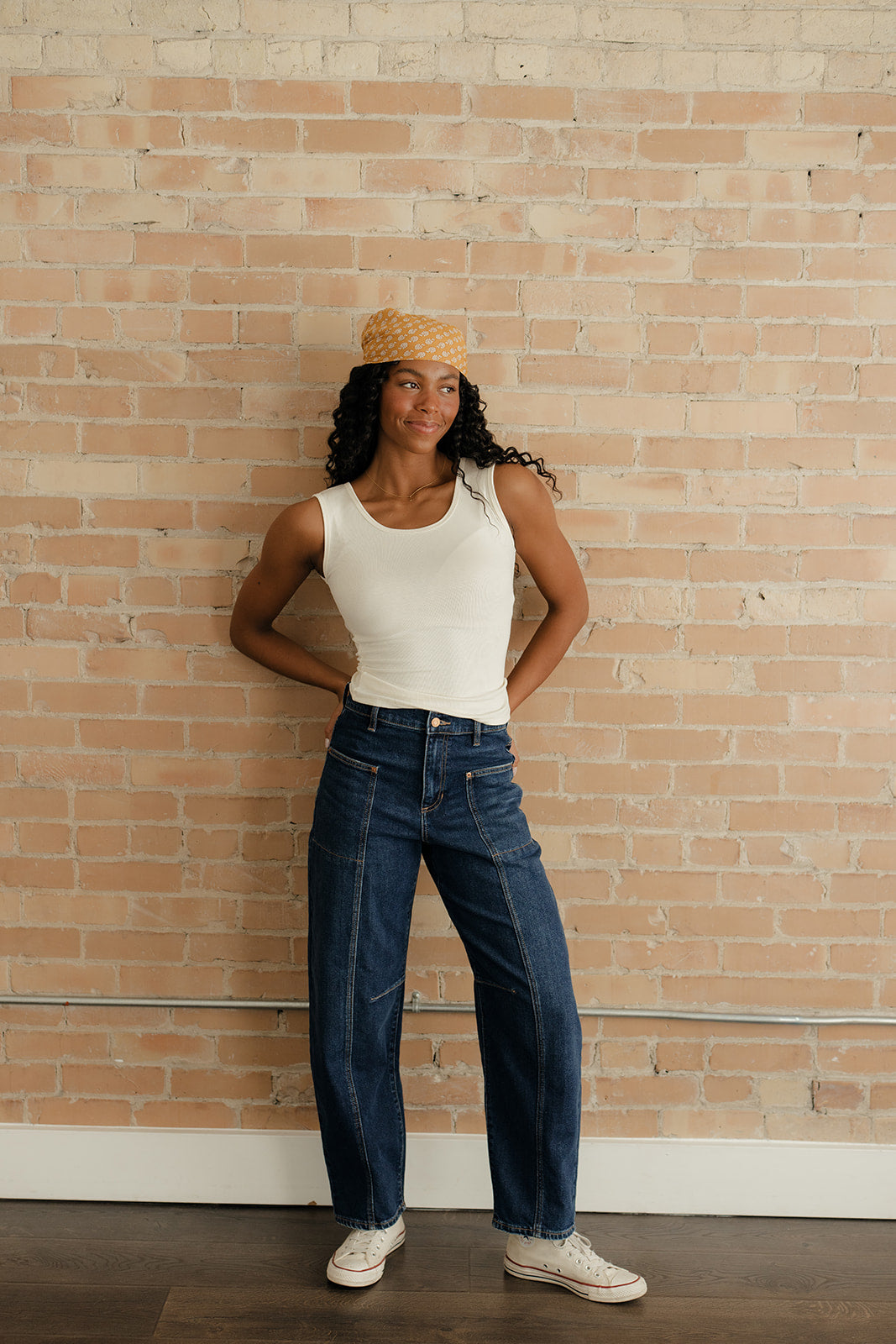 Woman wearing a white tank top and blue jeans standing against a brick wall.