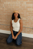 Woman kneeling in front of a brick wall wearing a white tank top and blue jeans.