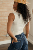 Woman wearing a white tank top and blue jeans with a yellow patterned headscarf, standing indoors.