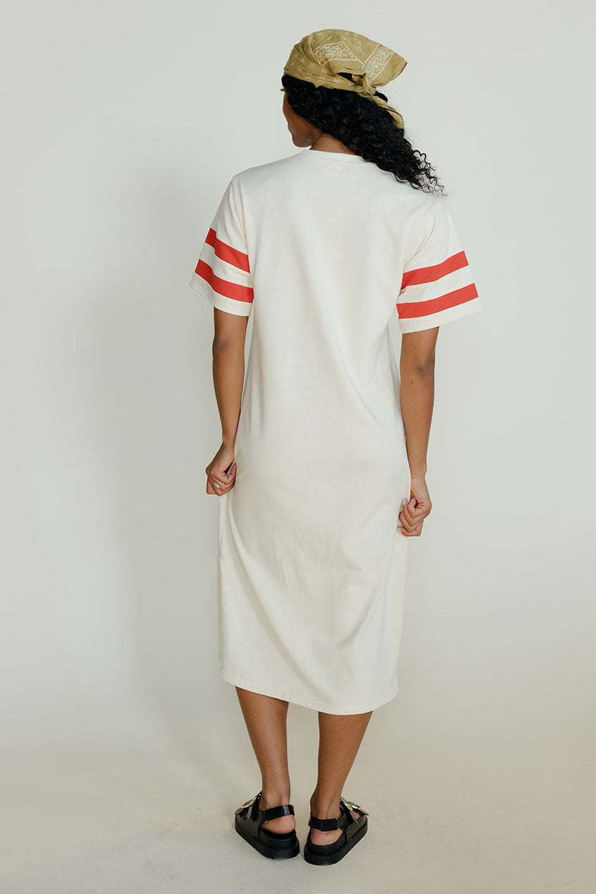 Person wearing a white dress with red stripes and a beige cap on a plain background