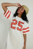 Woman wearing a white t-shirt with red numbers and a beige cap against a plain background