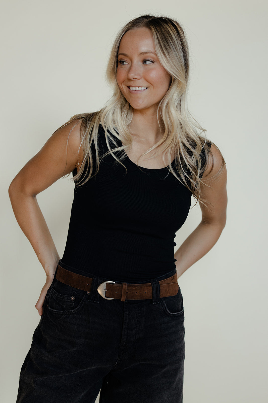 Woman wearing a black tank top and dark pants with a brown belt against a plain background