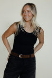 Woman wearing a black tank top and dark pants with a brown belt against a plain background