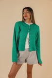 A woman wearing a green button-up cardigan with a rolled collar and arm cuffs, paired with light gray shorts.